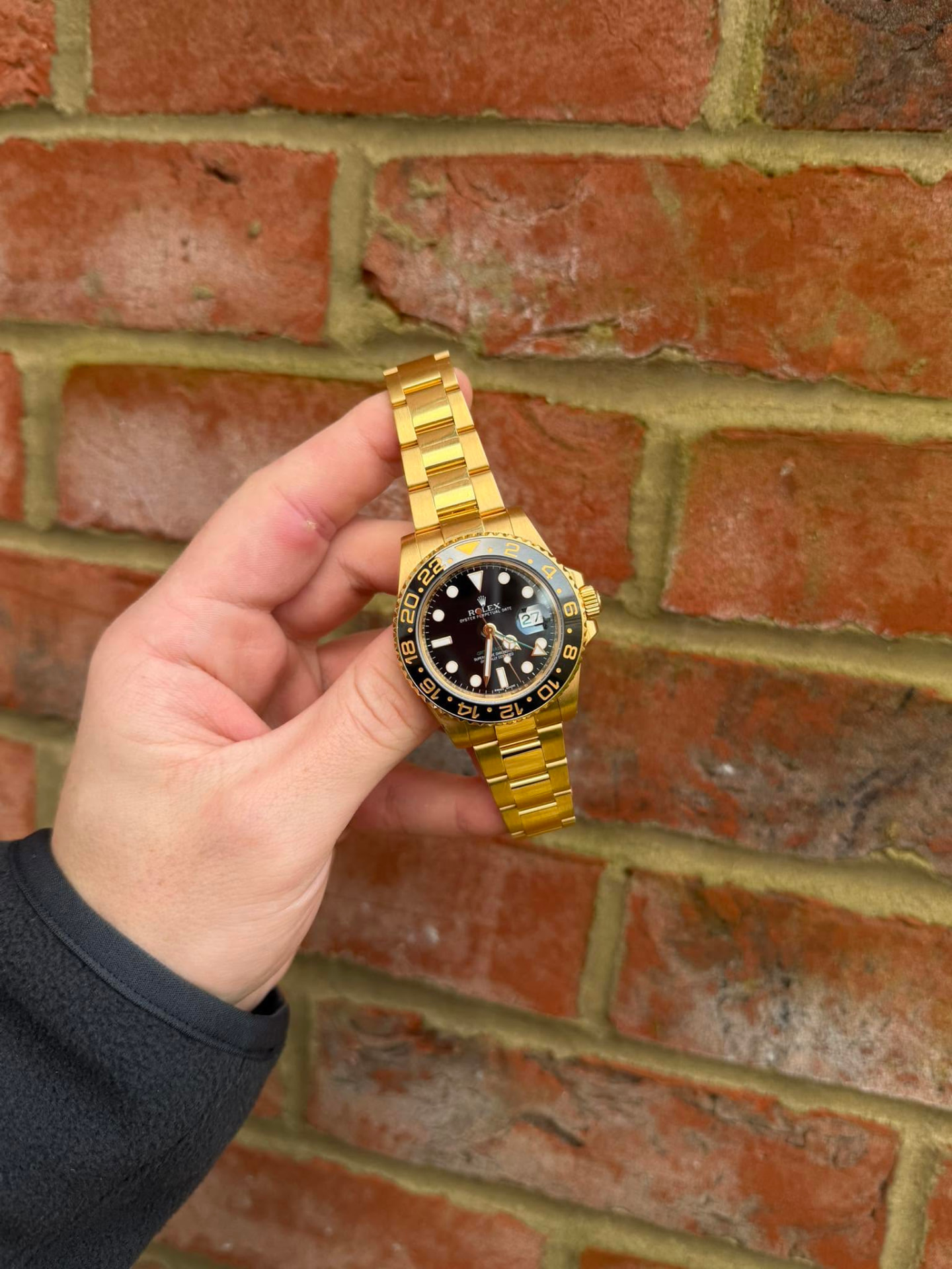 Gold luxury sports watch with a black dial held in front of a brick wall